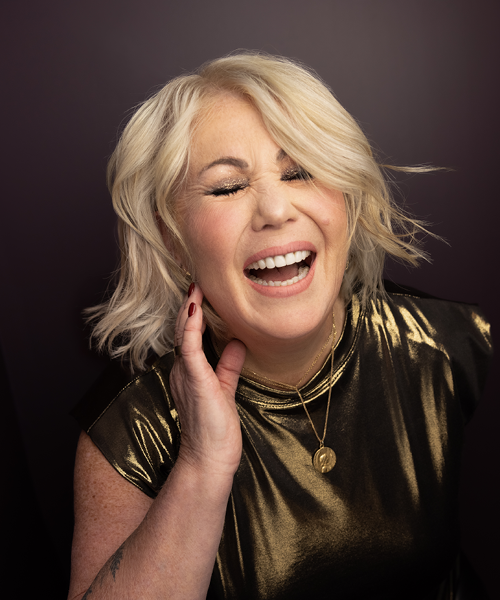 Jann Arden smiling with her eyes shut. She has blonde hair and wears a gold top. 